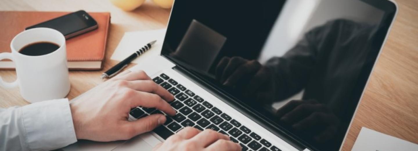 zwei Hände auf einer Laptop-Tastatur, daneben eine Kaffeetasse und Arbeitsutensilien
